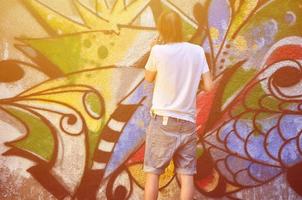 foto en el proceso de dibujar un patrón de graffiti en un antiguo muro de hormigón. un joven rubio de pelo largo dibuja un dibujo abstracto de diferentes colores. concepto de arte callejero y vandalismo