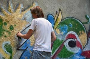 Photo in the process of drawing a graffiti pattern on an old concrete wall. Young long-haired blond guy draws an abstract drawing of different colors. Street art and vandalism concept