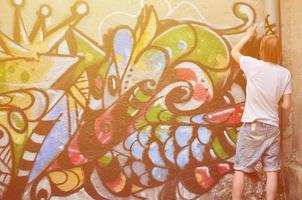 Photo in the process of drawing a graffiti pattern on an old concrete wall. Young long-haired blond guy draws an abstract drawing of different colors. Street art and vandalism concept