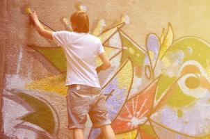 foto en el proceso de dibujar un patrón de graffiti en un antiguo muro de hormigón. un joven rubio de pelo largo dibuja un dibujo abstracto de diferentes colores. concepto de arte callejero y vandalismo