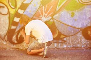 foto en el proceso de dibujar un patrón de graffiti en un antiguo muro de hormigón. un joven rubio de pelo largo dibuja un dibujo abstracto de diferentes colores. concepto de arte callejero y vandalismo