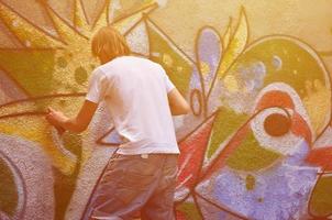 Photo of a young guy in denim shorts and a white shirt. The guy draws on the graffiti wall a drawing with aerosol paints of various colors. The concept of hooliganism and damage to property