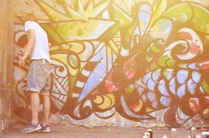 Photo of a young guy in denim shorts and a white shirt. The guy draws on the graffiti wall a drawing with aerosol paints of various colors. The concept of hooliganism and damage to property