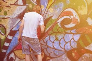 foto en el proceso de dibujar un patrón de graffiti en un antiguo muro de hormigón. un joven rubio de pelo largo dibuja un dibujo abstracto de diferentes colores. concepto de arte callejero y vandalismo