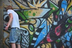 Photo in the process of drawing a graffiti pattern on an old concrete wall. Young long-haired blond guy draws an abstract drawing of different colors. Street art and vandalism concept