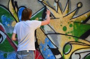 foto de un joven con pantalones cortos de mezclilla y una camisa blanca. el chico dibuja en la pared de graffiti un dibujo con pinturas en aerosol de varios colores. el concepto de vandalismo y daños a la propiedad