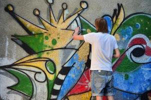 Photo of a young guy in denim shorts and a white shirt. The guy draws on the graffiti wall a drawing with aerosol paints of various colors. The concept of hooliganism and damage to property