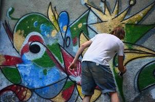 Photo of a young guy in denim shorts and a white shirt. The guy draws on the graffiti wall a drawing with aerosol paints of various colors. The concept of hooliganism and damage to property