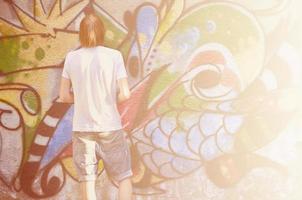 Photo of a young guy in denim shorts and a white shirt. The guy draws on the graffiti wall a drawing with aerosol paints of various colors. The concept of hooliganism and damage to property