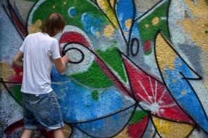 Photo in the process of drawing a graffiti pattern on an old concrete wall. Young long-haired blond guy draws an abstract drawing of different colors. Street art and vandalism concept