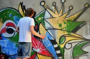 Photo of a young guy in denim shorts and a white shirt. The guy draws on the graffiti wall a drawing with aerosol paints of various colors. The concept of hooliganism and damage to property