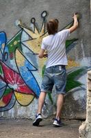 Photo in the process of drawing a graffiti pattern on an old concrete wall. Young long-haired blond guy draws an abstract drawing of different colors. Street art and vandalism concept