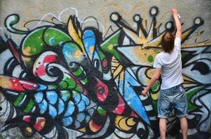 foto en el proceso de dibujar un patrón de graffiti en un antiguo muro de hormigón. un joven rubio de pelo largo dibuja un dibujo abstracto de diferentes colores. concepto de arte callejero y vandalismo