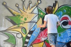 foto en el proceso de dibujar un patrón de graffiti en un antiguo muro de hormigón. un joven rubio de pelo largo dibuja un dibujo abstracto de diferentes colores. concepto de arte callejero y vandalismo