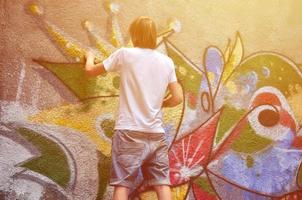 Photo of a young guy in denim shorts and a white shirt. The guy draws on the graffiti wall a drawing with aerosol paints of various colors. The concept of hooliganism and damage to property