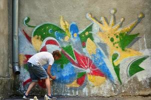 Photo of a young guy in denim shorts and a white shirt. The guy draws on the graffiti wall a drawing with aerosol paints of various colors. The concept of hooliganism and damage to property