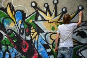 Photo of a young guy in denim shorts and a white shirt. The guy draws on the graffiti wall a drawing with aerosol paints of various colors. The concept of hooliganism and damage to property