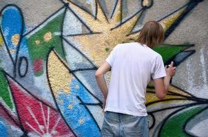 foto en el proceso de dibujar un patrón de graffiti en un antiguo muro de hormigón. un joven rubio de pelo largo dibuja un dibujo abstracto de diferentes colores. concepto de arte callejero y vandalismo