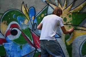 Photo of a young guy in denim shorts and a white shirt. The guy draws on the graffiti wall a drawing with aerosol paints of various colors. The concept of hooliganism and damage to property