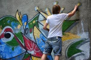 foto en el proceso de dibujar un patrón de graffiti en un antiguo muro de hormigón. un joven rubio de pelo largo dibuja un dibujo abstracto de diferentes colores. concepto de arte callejero y vandalismo