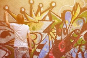 Photo of a young guy in denim shorts and a white shirt. The guy draws on the graffiti wall a drawing with aerosol paints of various colors. The concept of hooliganism and damage to property