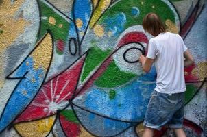 foto en el proceso de dibujar un patrón de graffiti en un antiguo muro de hormigón. un joven rubio de pelo largo dibuja un dibujo abstracto de diferentes colores. concepto de arte callejero y vandalismo