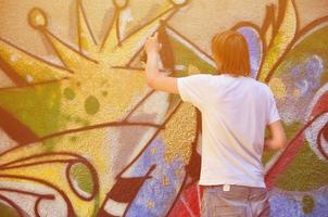 Photo of a young guy in denim shorts and a white shirt. The guy draws on the graffiti wall a drawing with aerosol paints of various colors. The concept of hooliganism and damage to property