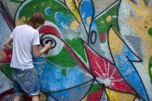 Photo of a young guy in denim shorts and a white shirt. The guy draws on the graffiti wall a drawing with aerosol paints of various colors. The concept of hooliganism and damage to property