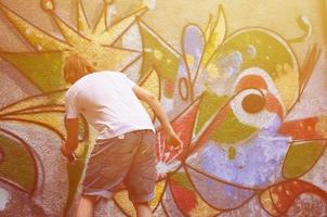 Photo of a young guy in denim shorts and a white shirt. The guy draws on the graffiti wall a drawing with aerosol paints of various colors. The concept of hooliganism and damage to property
