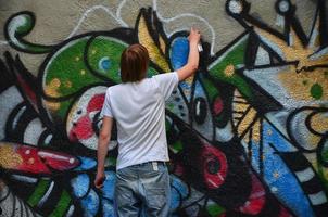 foto en el proceso de dibujar un patrón de graffiti en un antiguo muro de hormigón. un joven rubio de pelo largo dibuja un dibujo abstracto de diferentes colores. concepto de arte callejero y vandalismo