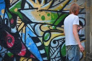 foto en el proceso de dibujar un patrón de graffiti en un antiguo muro de hormigón. un joven rubio de pelo largo dibuja un dibujo abstracto de diferentes colores. concepto de arte callejero y vandalismo