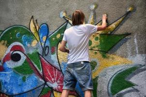 Photo of a young guy in denim shorts and a white shirt. The guy draws on the graffiti wall a drawing with aerosol paints of various colors. The concept of hooliganism and damage to property