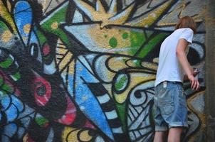 Photo in the process of drawing a graffiti pattern on an old concrete wall. Young long-haired blond guy draws an abstract drawing of different colors. Street art and vandalism concept