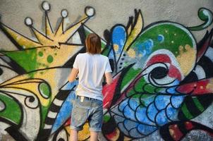 foto en el proceso de dibujar un patrón de graffiti en un antiguo muro de hormigón. un joven rubio de pelo largo dibuja un dibujo abstracto de diferentes colores. concepto de arte callejero y vandalismo