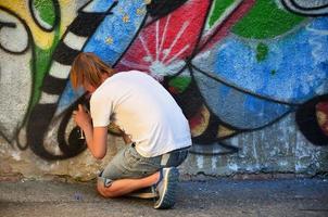 foto en el proceso de dibujar un patrón de graffiti en un antiguo muro de hormigón. un joven rubio de pelo largo dibuja un dibujo abstracto de diferentes colores. concepto de arte callejero y vandalismo