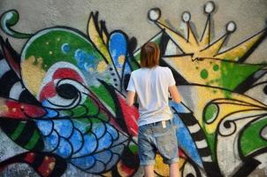 Photo in the process of drawing a graffiti pattern on an old concrete wall. Young long-haired blond guy draws an abstract drawing of different colors. Street art and vandalism concept