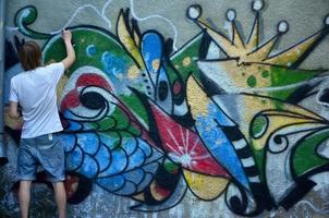 Photo of a young guy in denim shorts and a white shirt. The guy draws on the graffiti wall a drawing with aerosol paints of various colors. The concept of hooliganism and damage to property