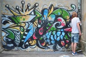 Photo in the process of drawing a graffiti pattern on an old concrete wall. Young long-haired blond guy draws an abstract drawing of different colors. Street art and vandalism concept