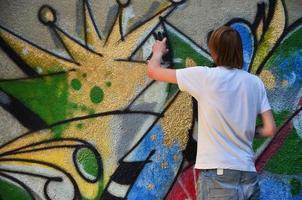 foto en el proceso de dibujar un patrón de graffiti en un antiguo muro de hormigón. un joven rubio de pelo largo dibuja un dibujo abstracto de diferentes colores. concepto de arte callejero y vandalismo