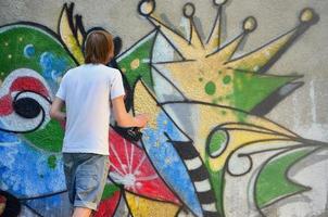 foto en el proceso de dibujar un patrón de graffiti en un antiguo muro de hormigón. un joven rubio de pelo largo dibuja un dibujo abstracto de diferentes colores. concepto de arte callejero y vandalismo