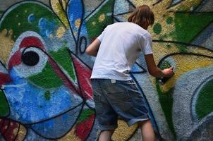 foto en el proceso de dibujar un patrón de graffiti en un antiguo muro de hormigón. un joven rubio de pelo largo dibuja un dibujo abstracto de diferentes colores. concepto de arte callejero y vandalismo