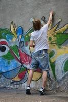 foto en el proceso de dibujar un patrón de graffiti en un antiguo muro de hormigón. un joven rubio de pelo largo dibuja un dibujo abstracto de diferentes colores. concepto de arte callejero y vandalismo