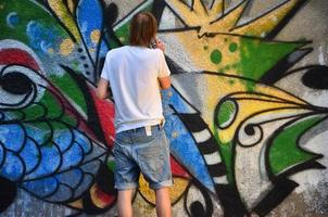 foto en el proceso de dibujar un patrón de graffiti en un antiguo muro de hormigón. un joven rubio de pelo largo dibuja un dibujo abstracto de diferentes colores. concepto de arte callejero y vandalismo