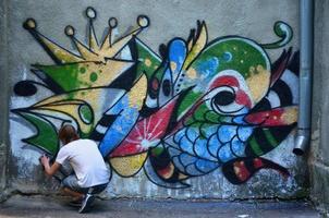 foto en el proceso de dibujar un patrón de graffiti en un antiguo muro de hormigón. un joven rubio de pelo largo dibuja un dibujo abstracto de diferentes colores. concepto de arte callejero y vandalismo