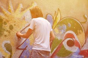 foto en el proceso de dibujar un patrón de graffiti en un antiguo muro de hormigón. un joven rubio de pelo largo dibuja un dibujo abstracto de diferentes colores. concepto de arte callejero y vandalismo