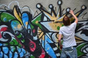 Photo in the process of drawing a graffiti pattern on an old concrete wall. Young long-haired blond guy draws an abstract drawing of different colors. Street art and vandalism concept