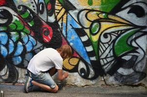 Photo in the process of drawing a graffiti pattern on an old concrete wall. Young long-haired blond guy draws an abstract drawing of different colors. Street art and vandalism concept