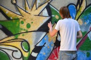 foto en el proceso de dibujar un patrón de graffiti en un antiguo muro de hormigón. un joven rubio de pelo largo dibuja un dibujo abstracto de diferentes colores. concepto de arte callejero y vandalismo