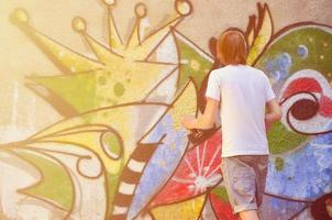 foto en el proceso de dibujar un patrón de graffiti en un antiguo muro de hormigón. un joven rubio de pelo largo dibuja un dibujo abstracto de diferentes colores. concepto de arte callejero y vandalismo