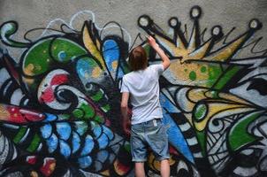 Photo of a young guy in denim shorts and a white shirt. The guy draws on the graffiti wall a drawing with aerosol paints of various colors. The concept of hooliganism and damage to property